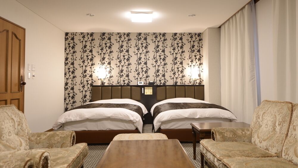 Una habitación de hotel con dos camas, una puerta de madera, un papel tapiz estampado y una pequeña mesa.