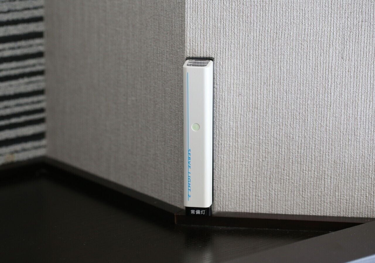 Un dispositivo blanco con una franja azul y una luz verde, etiquetado como "SERVE LIGHT 2", montado en una pared texturizada cerca de una superficie negra.