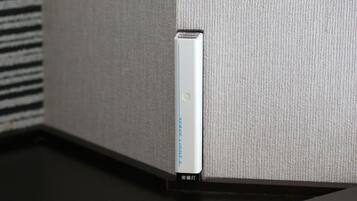 Un dispositivo blanco con una franja azul y una luz verde, etiquetado como "SERVE LIGHT 2", montado en una pared texturizada cerca de una superficie negra.