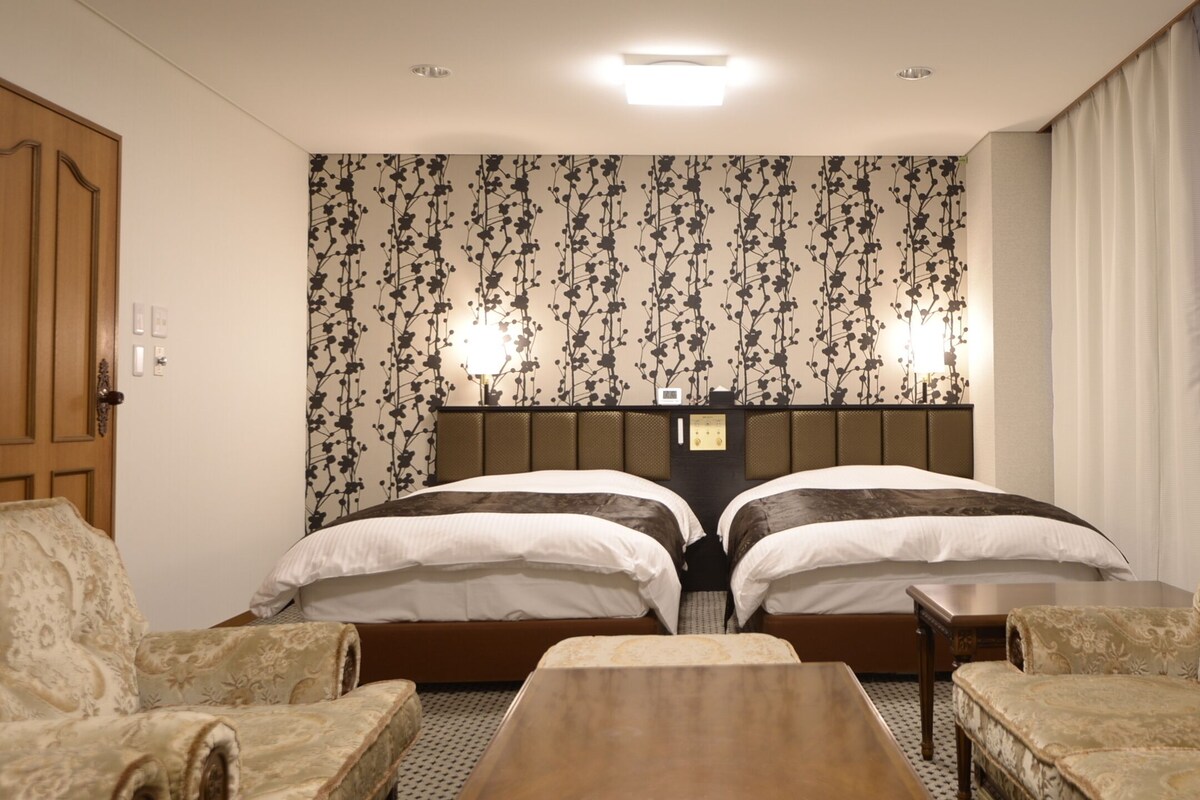 Une chambre d’hôtel avec deux lits, une tête de lit en bois, une petite table, deux fauteuils et un papier peint à motifs.