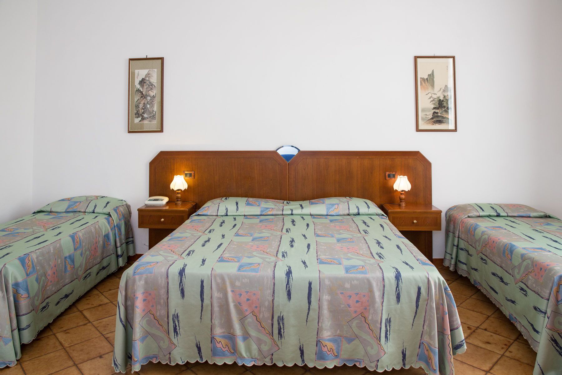 Habitación de hotel con cabecera de madera, dos mesitas de noche con lámparas, cama con ropa de cama estampada y dos cuadros en las paredes.