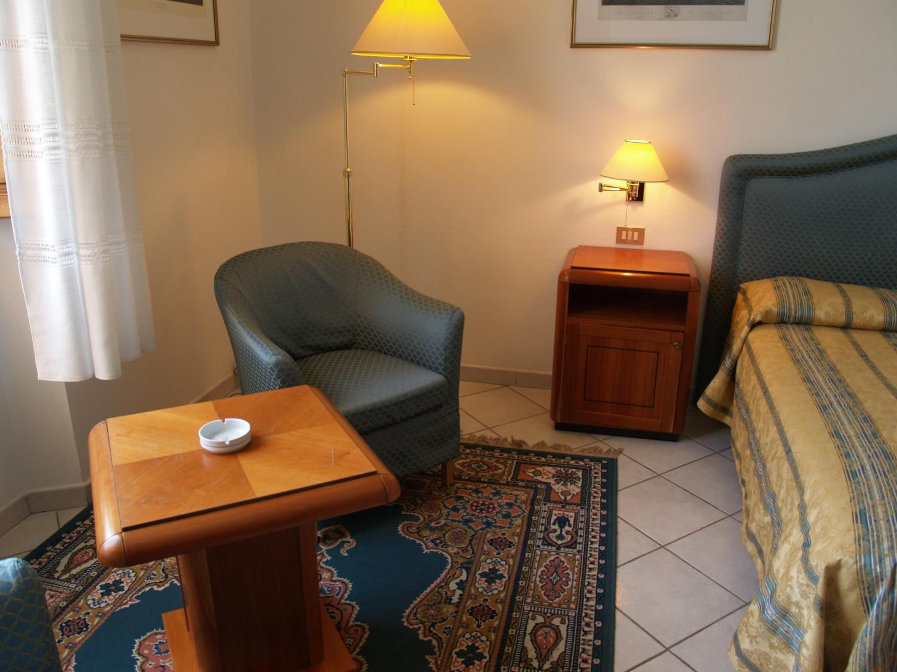 A hotel room with a bed, a chair, a nightstand, a table, and a patterned rug.