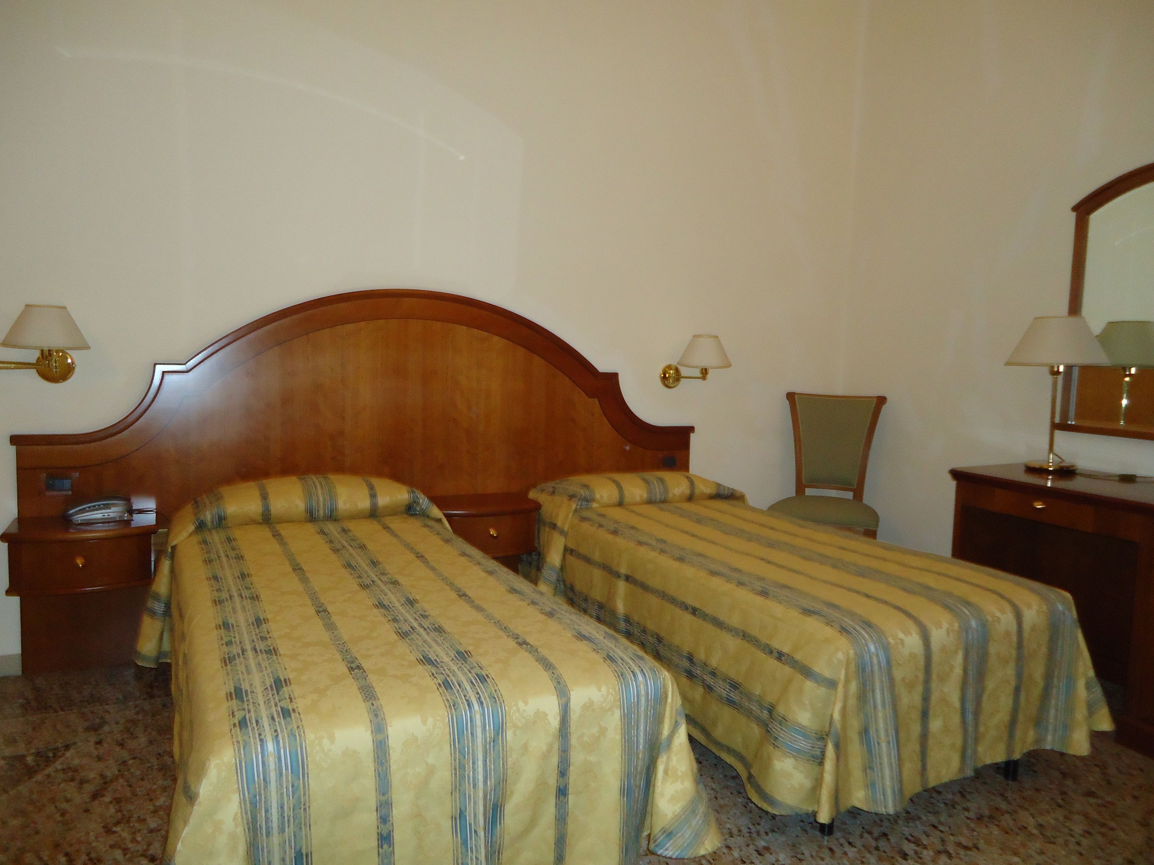 A hotel room with two single beds, wooden headboards, bedside tables with lamps, a chair, and a dresser.