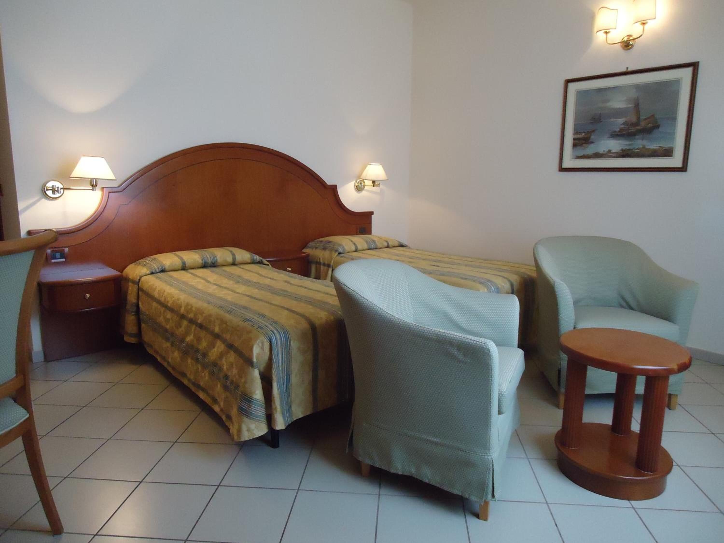 Habitación de hotel con dos camas, una silla, una mesita y un cuadro en la pared.