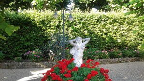 En statue af en kvinde med løftede arme, omgivet af et bed med røde blomster, foran et væld af grønne blade og en stenvæg.