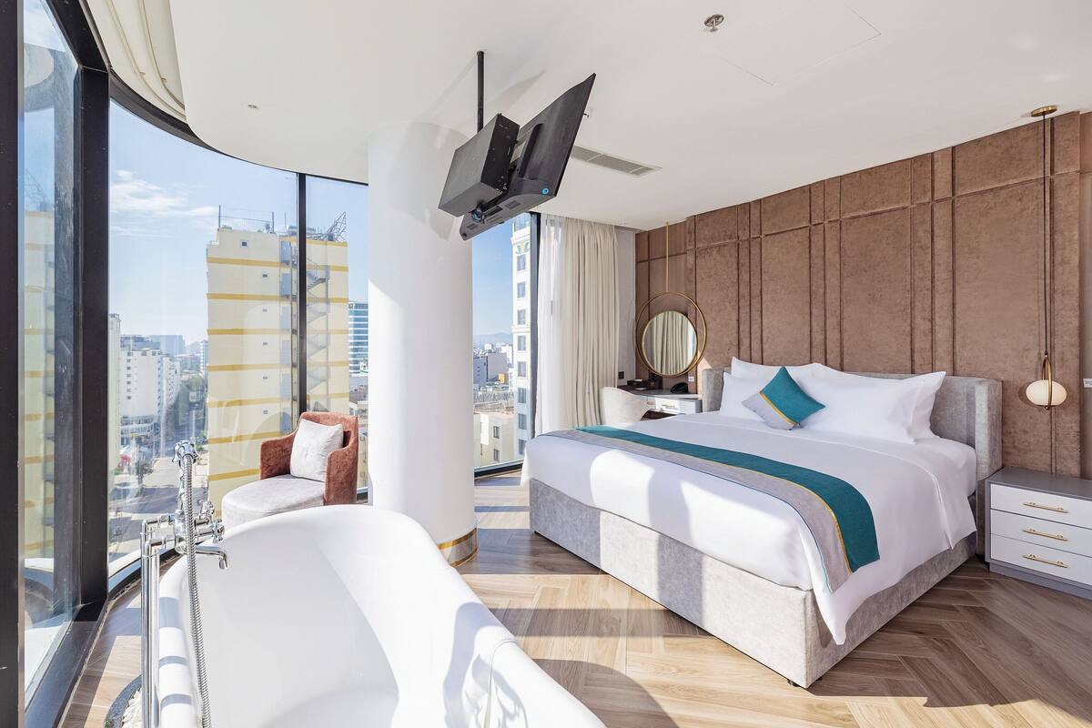 Een moderne hotelkamer met een groot bed, een zithoek met een fauteuil, een badkuip, een wastafel met spiegel en een aan het plafond bevestigde televisie.