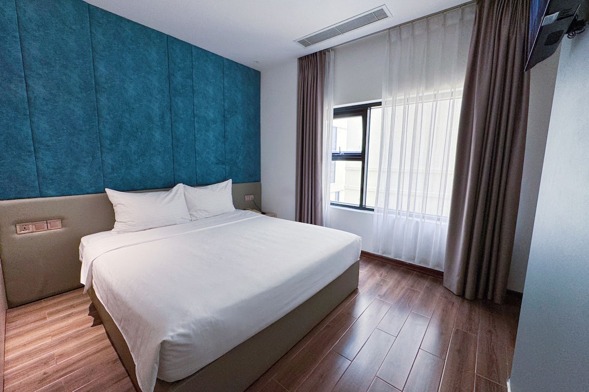 Hotelkamer met een groot bed, hoofdbord, raam met gordijnen en een wand met een textuurpaneel.