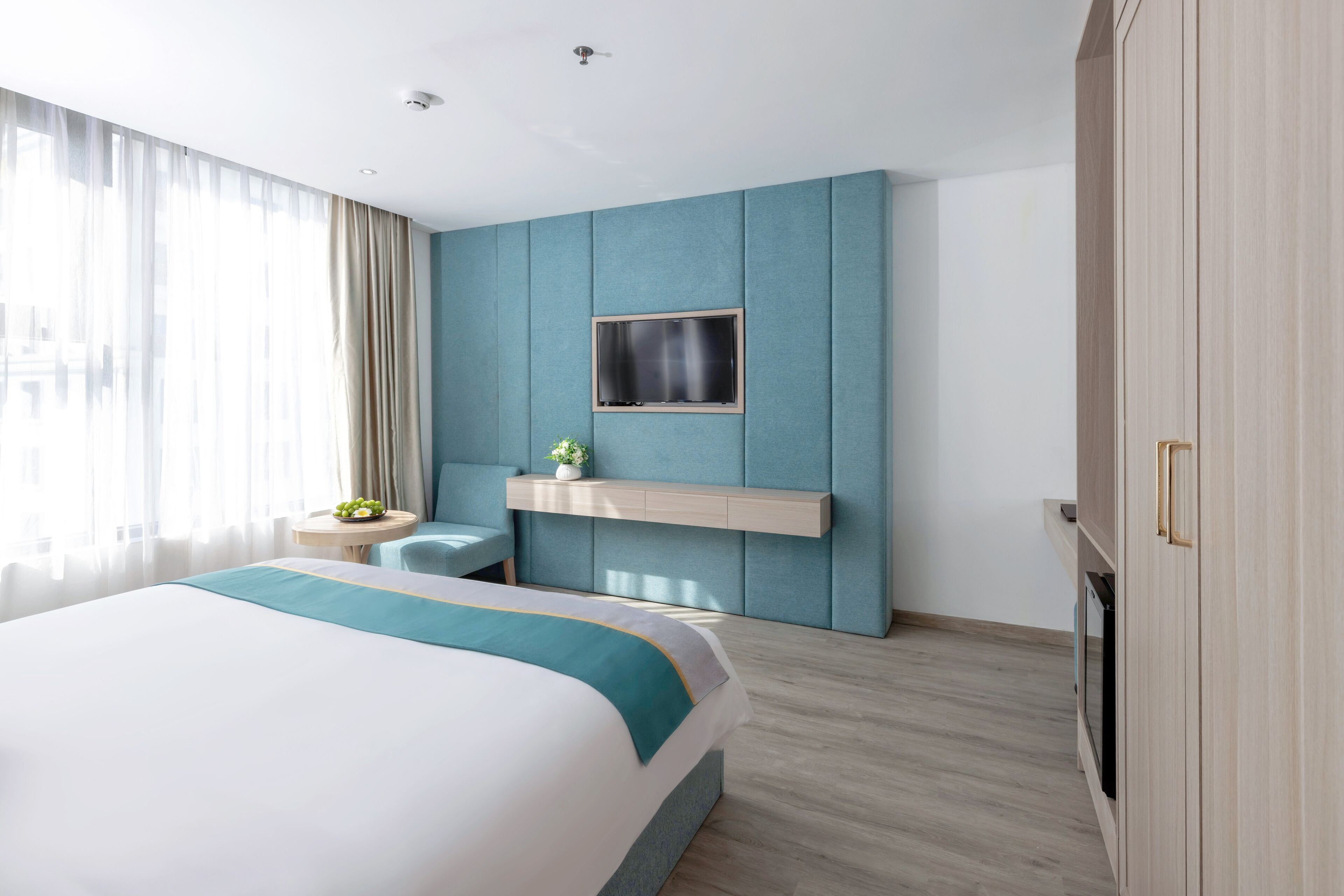 A modern hotel room with a large bed, a flat-screen TV mounted on a blue panel, a small round table with a vase of flowers, and a wardrobe with a gold handle.