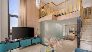 Uma sala moderna com uma grande TV de tela plana, painel de entretenimento preto e verde-água, mesa de centro de vidro com plantas decorativas, e vista para a cozinha e banheiro através de uma divisória de vidro.
