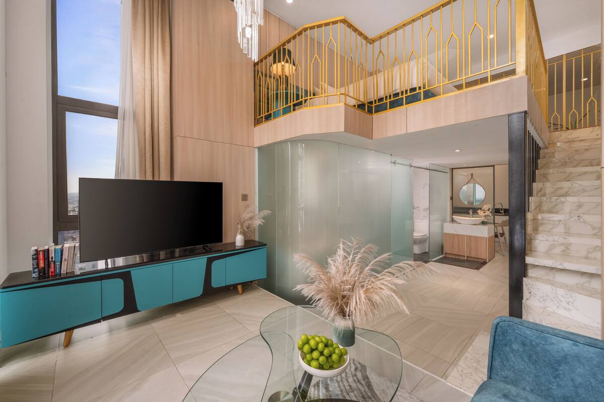 Een moderne woonkamer met een grote flatscreen-tv, een entertainmentmeubel in teal en zwart, een glazen salontafel met decoratieve planten, en een uitzicht op de keuken en badkamer door een glazen scheidingswand.