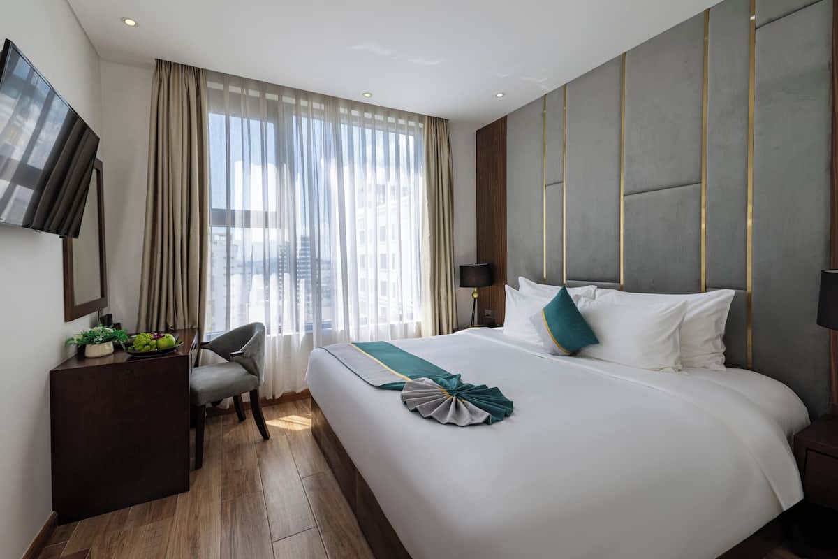 Een hotelkamer met een groot bed, een bureau met een flatscreen televisie, een stoel en uitzicht op de stad door dunne gordijnen.