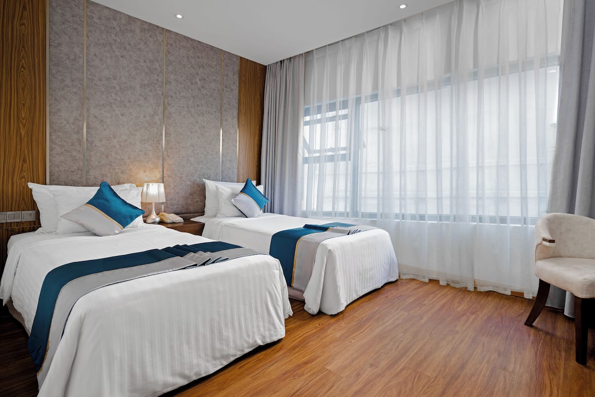 Een hotelkamer met twee bedden, een houten vloer, een groot raam met dunne gordijnen en een stoel.