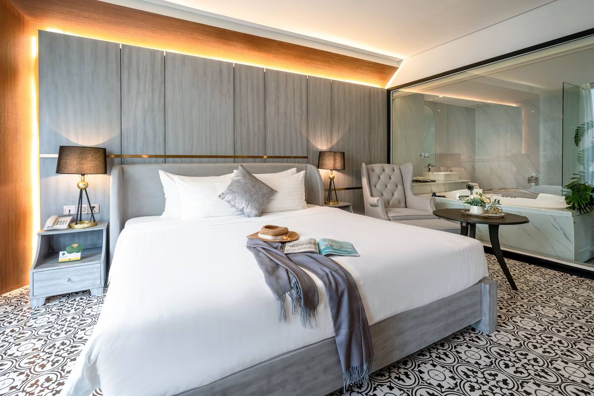 Een moderne hotelkamer met een groot bed, twee nachtkastjes met lampen, een stoel, een klein tafeltje met een plant, en een badkamer met bad en wastafel.