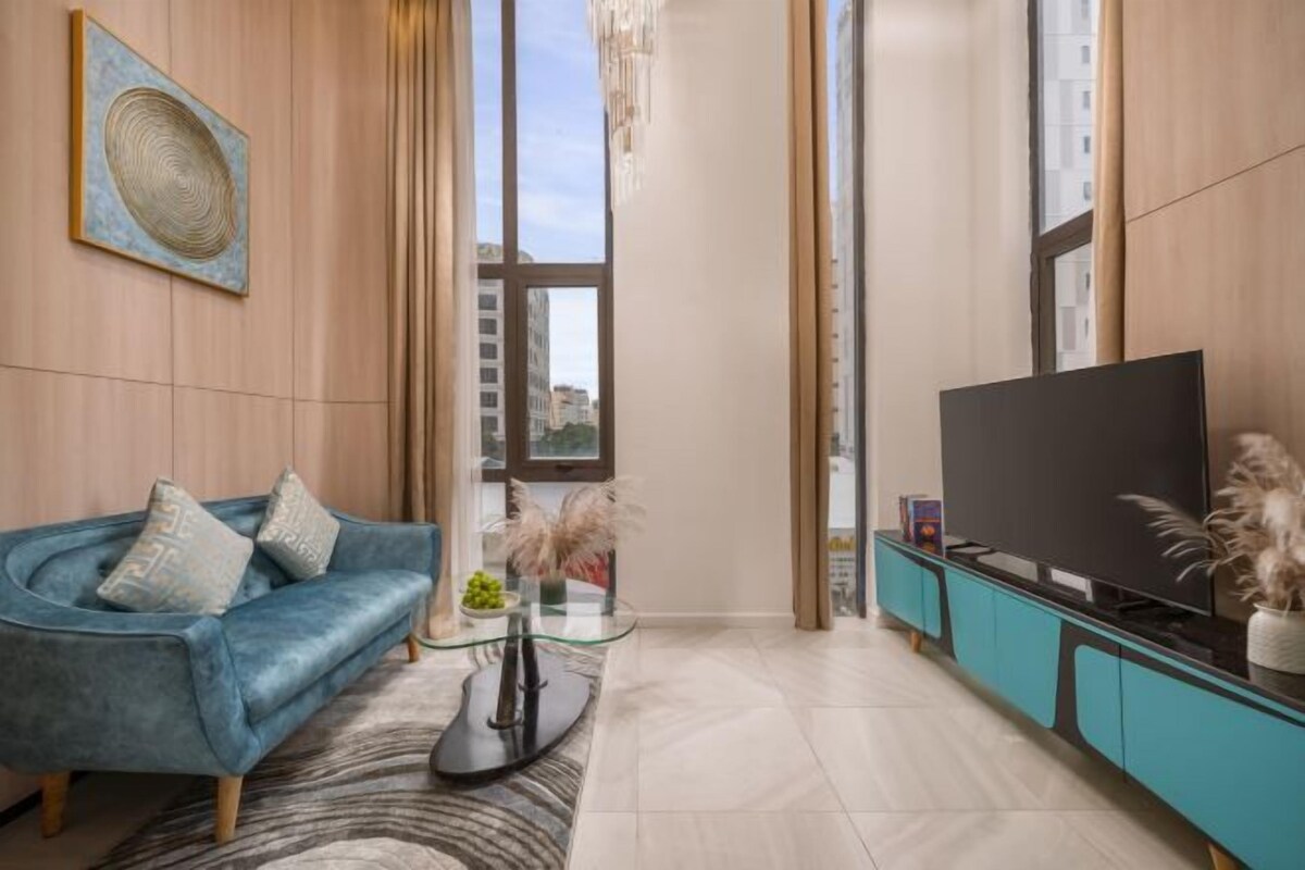 Un salon moderne avec un canapé bleu, une table basse en verre, un téléviseur à écran plat posé sur un meuble turquoise et une grande fenêtre donnant sur la ville.