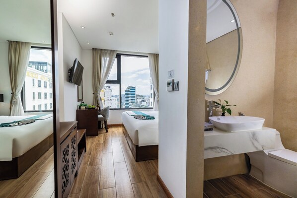 A hotel room with a bed, a desk, a chair, a TV, a window with curtains, and a bathroom with a sink and mirror. - Cicilia Hotels & Spa Danang (Da Nang)