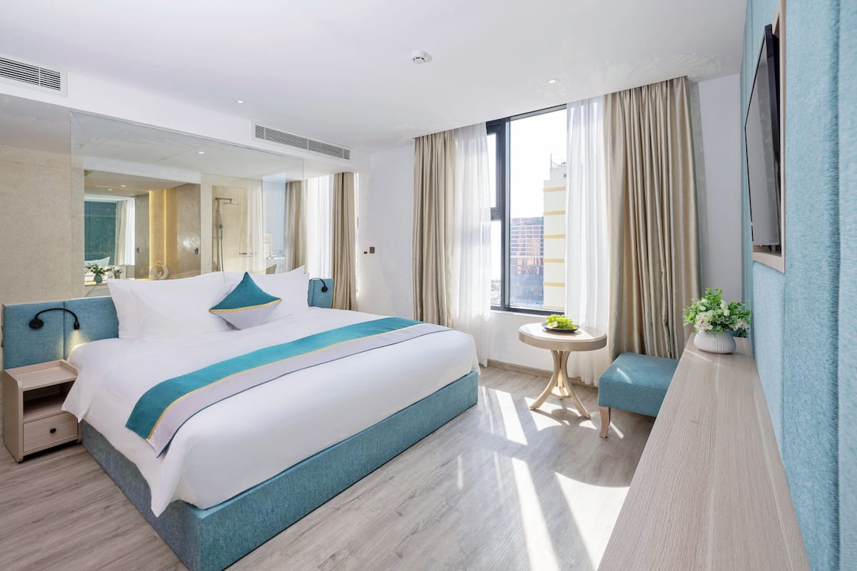 Een moderne hotelkamer met een groot bed, een nachtkastje, een kleine ronde tafel met een plant en een groot raam met gordijnen.