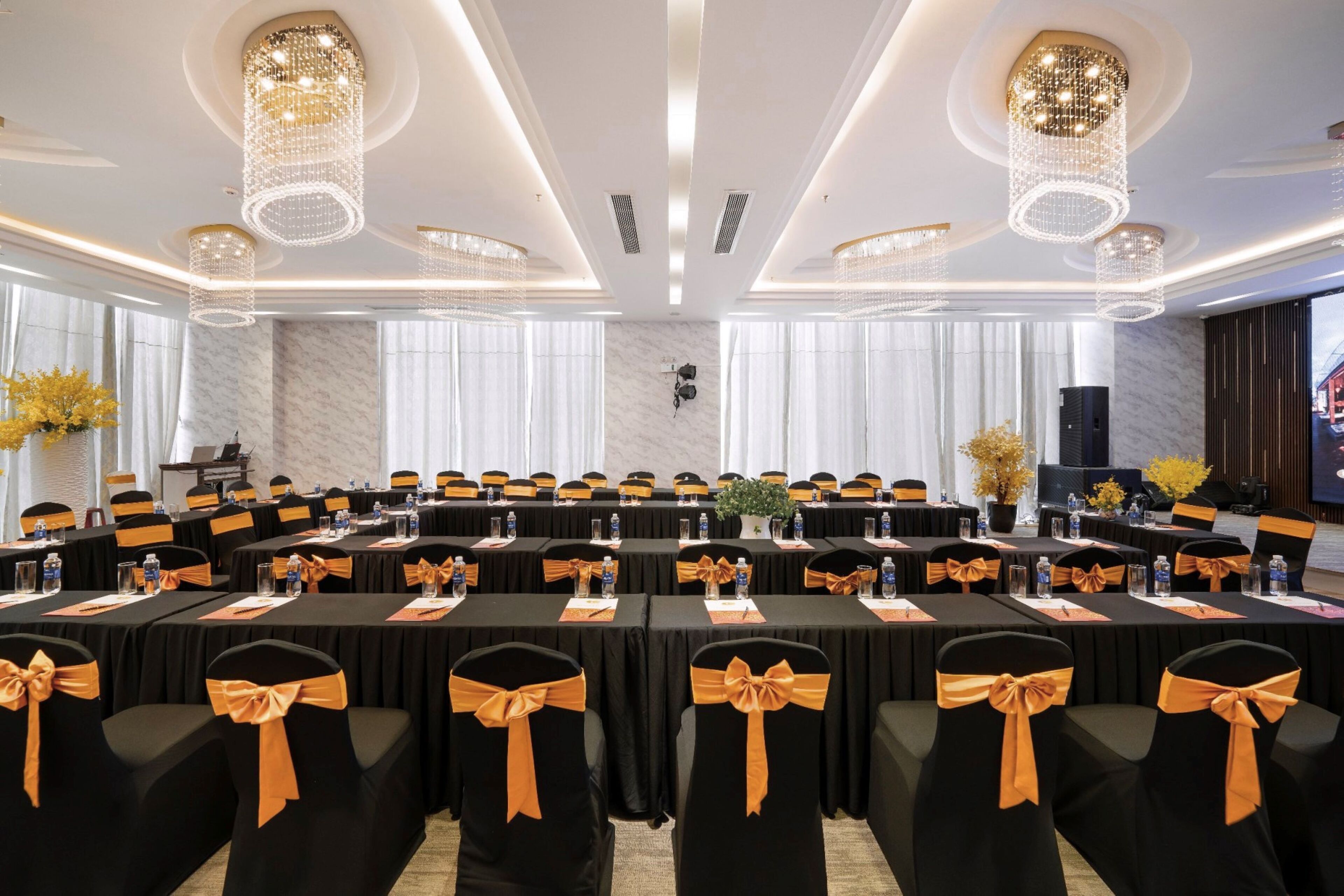 Une salle de conférence avec des tables rondes, des chaises noires ornées de rubans orange et des lustres.