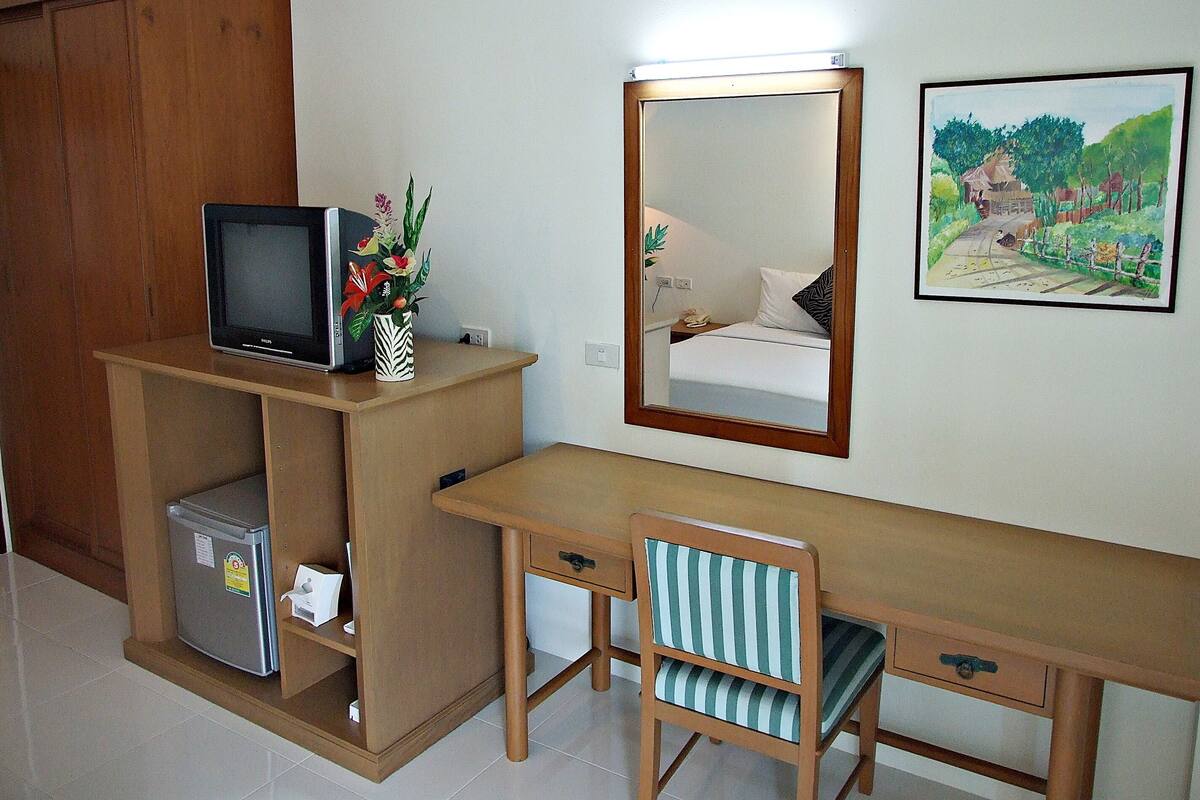 A compact bedroom with a wooden desk, a chair, a bed, a mirror, and a small refrigerator.