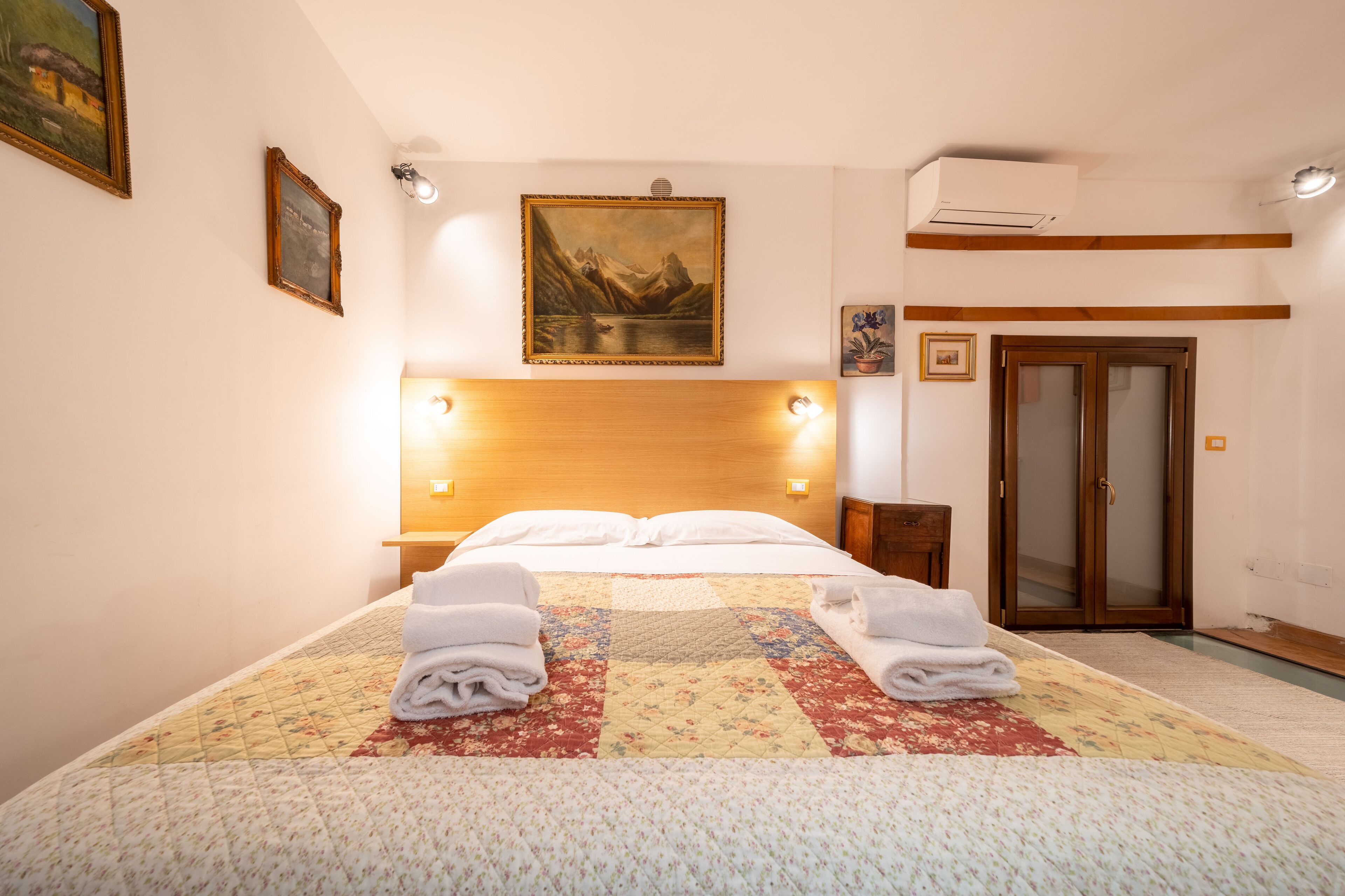 A neatly made bed with a quilt and towels, a wooden headboard, a painting above, and a sliding door to the right.
