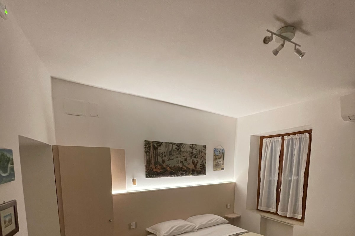 Una camera d'albergo con un letto, un tavolino, una sedia, un armadio, una ventola a soffitto e una finestra con tende.