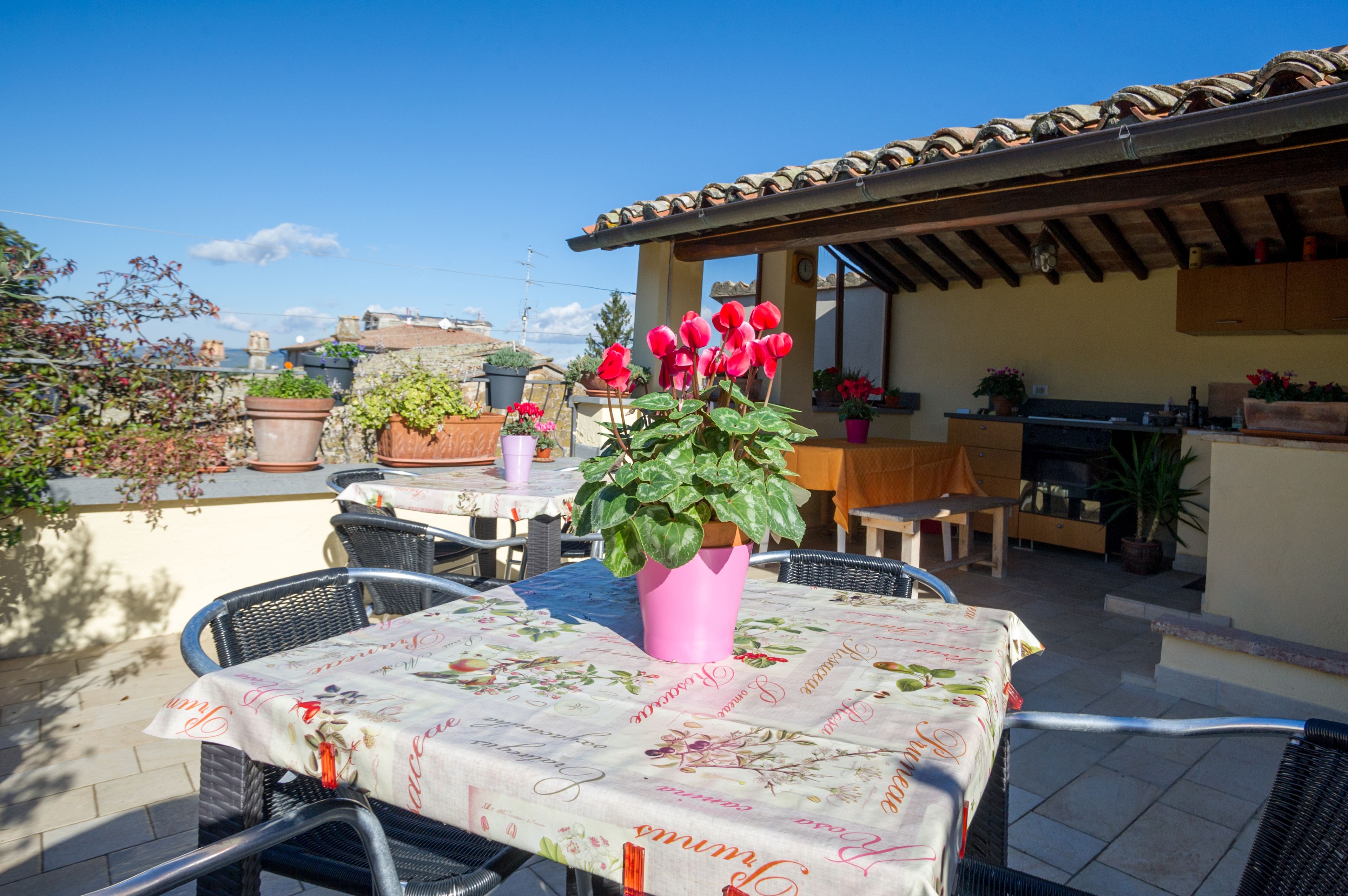 Un patio exterior con mesa lista para comer, rodeado de plantas en macetas y una estructura de pérgola.