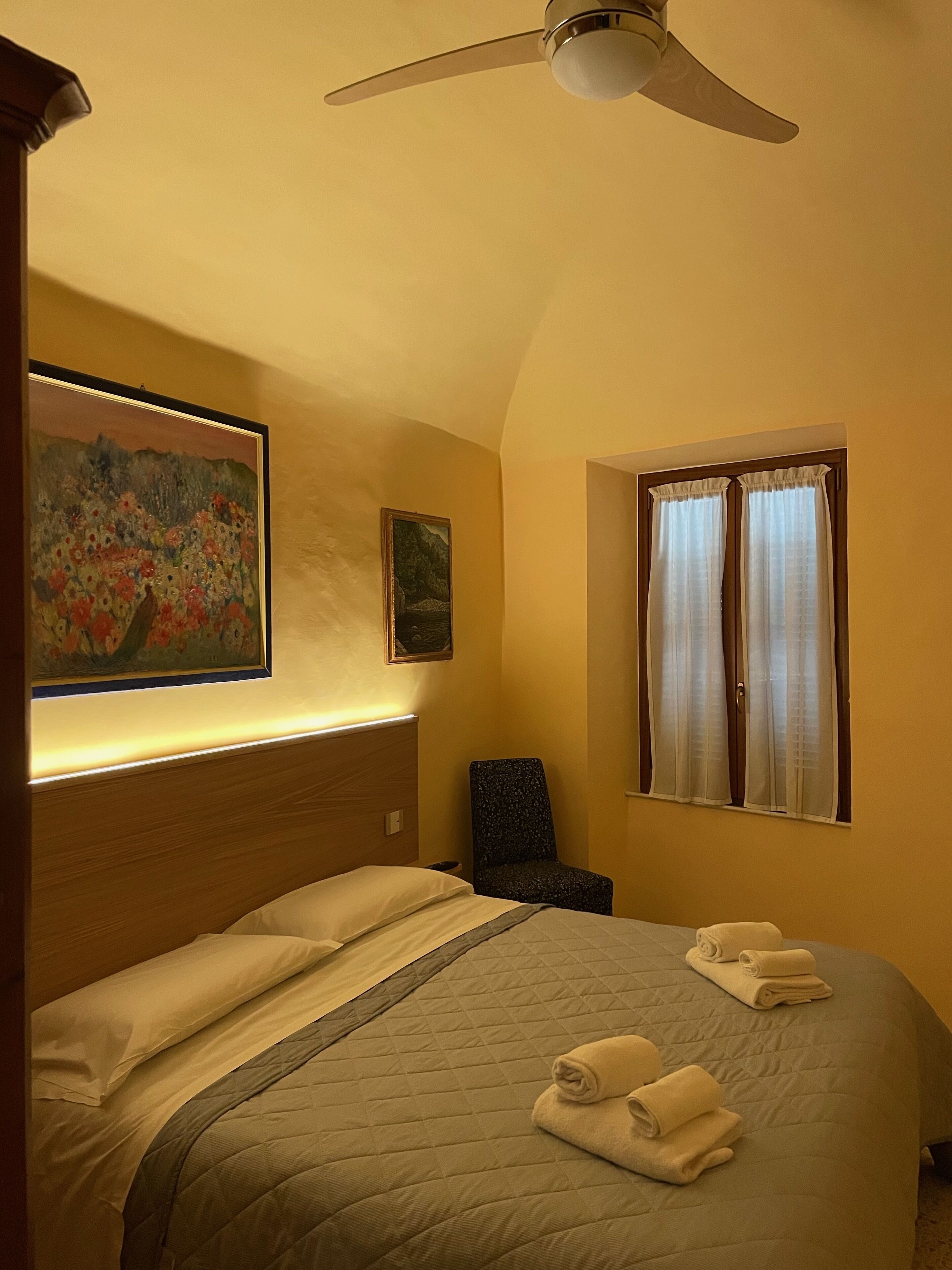 A hotel room with a bed, a chair, a ceiling fan, and two windows with curtains.