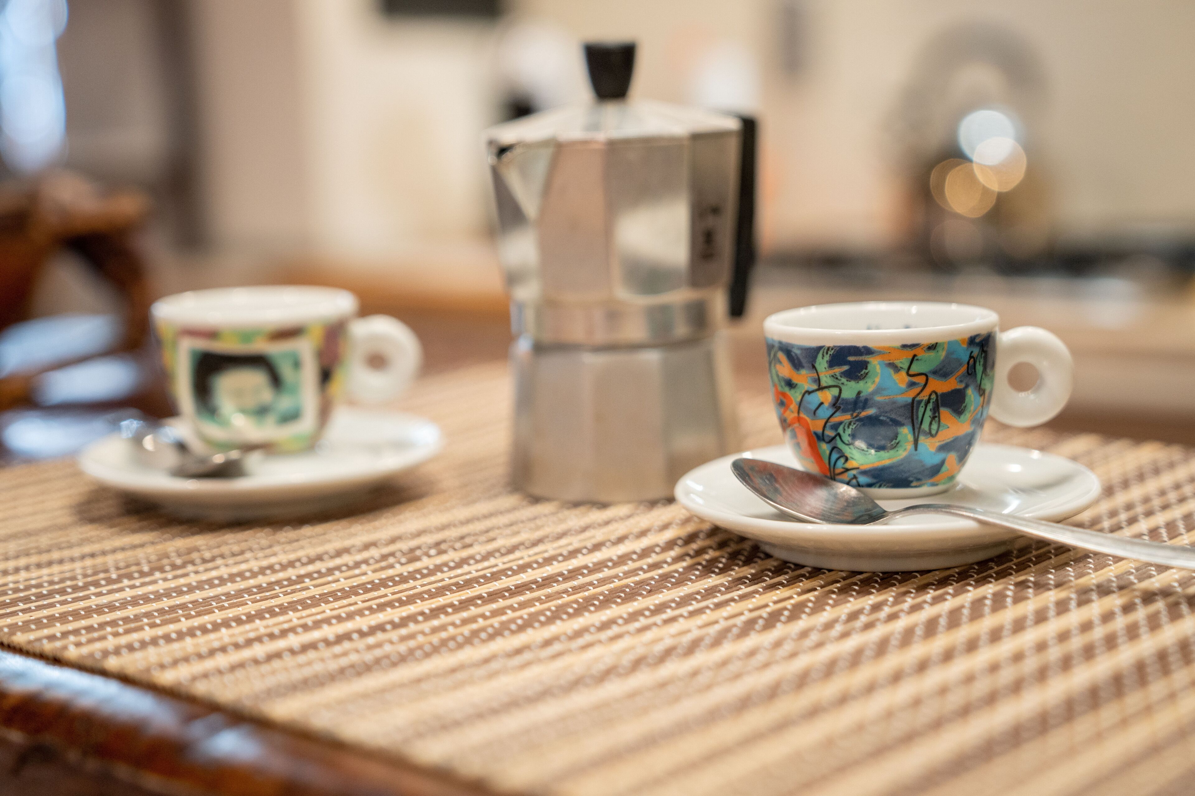 Two cups on saucers with spoons, a metallic coffee pot, and a textured placemat on a wooden table.