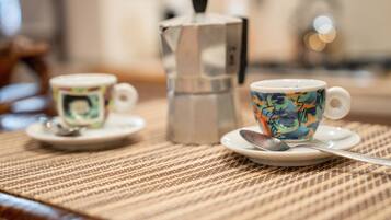 Two cups on saucers with spoons, a metallic coffee pot, and a textured placemat on a wooden table.