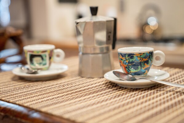 Two cups on saucers with spoons, a metallic coffee pot, and a textured placemat on a wooden table.