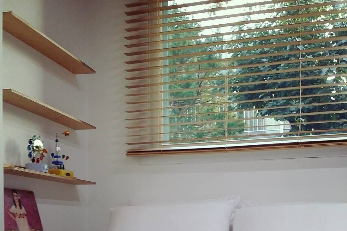 Une chambre à coucher avec un lit, des oreillers blancs, une couverture foncée, des étagères en bois et une fenêtre avec des stores.