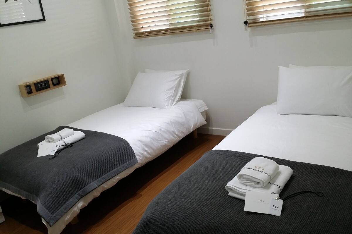 Une chambre d’hôtel avec deux lits simples, chacun recouvert de draps blancs et d’une couverture gris foncé. Les fenêtres sont équipées de stores en bois, et un petit étagère avec un tableau encadré et quelques objets est fixée au mur.
