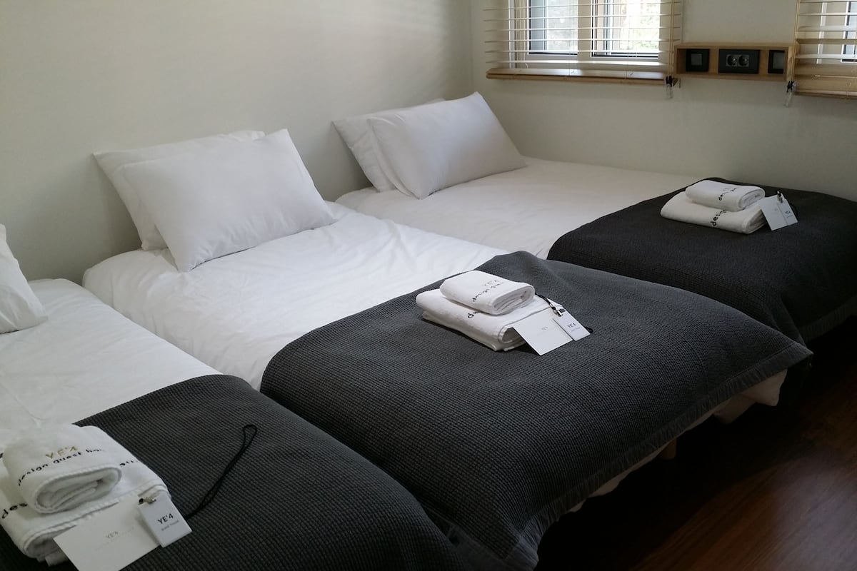 Une chambre d’hôtel avec trois lits, chacun étant équipé de draps blancs et d’une couverture gris foncé. Des serviettes, encore avec leurs étiquettes, sont posées sur les lits.