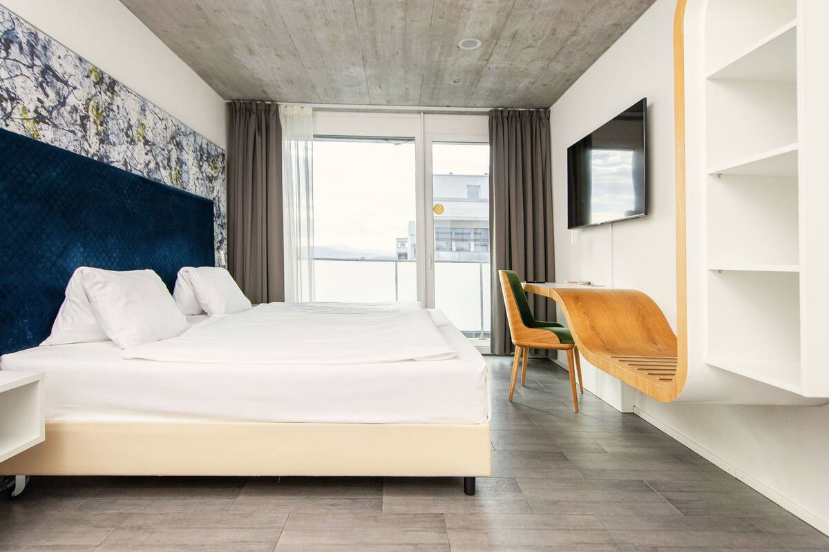 Una habitación de hotel moderna con una cama grande, un televisor montado en la pared y un balcón con vistas.
