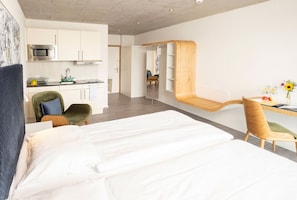 A modern hotel room with a bed, a desk with a chair, a kitchenette, and a bathroom.