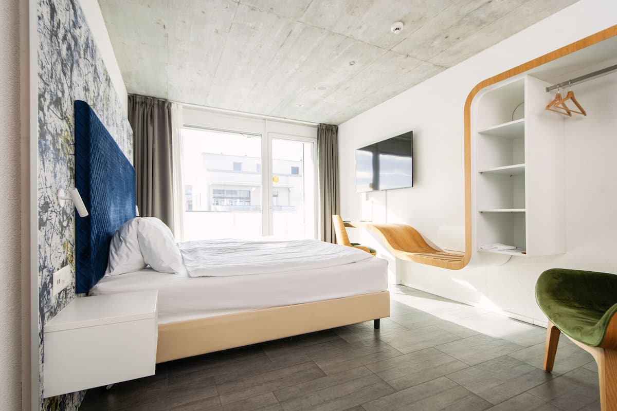 Una habitación de hotel moderna con una cama, un televisor montado en la pared y una zona de estar con una silla y una mesita.