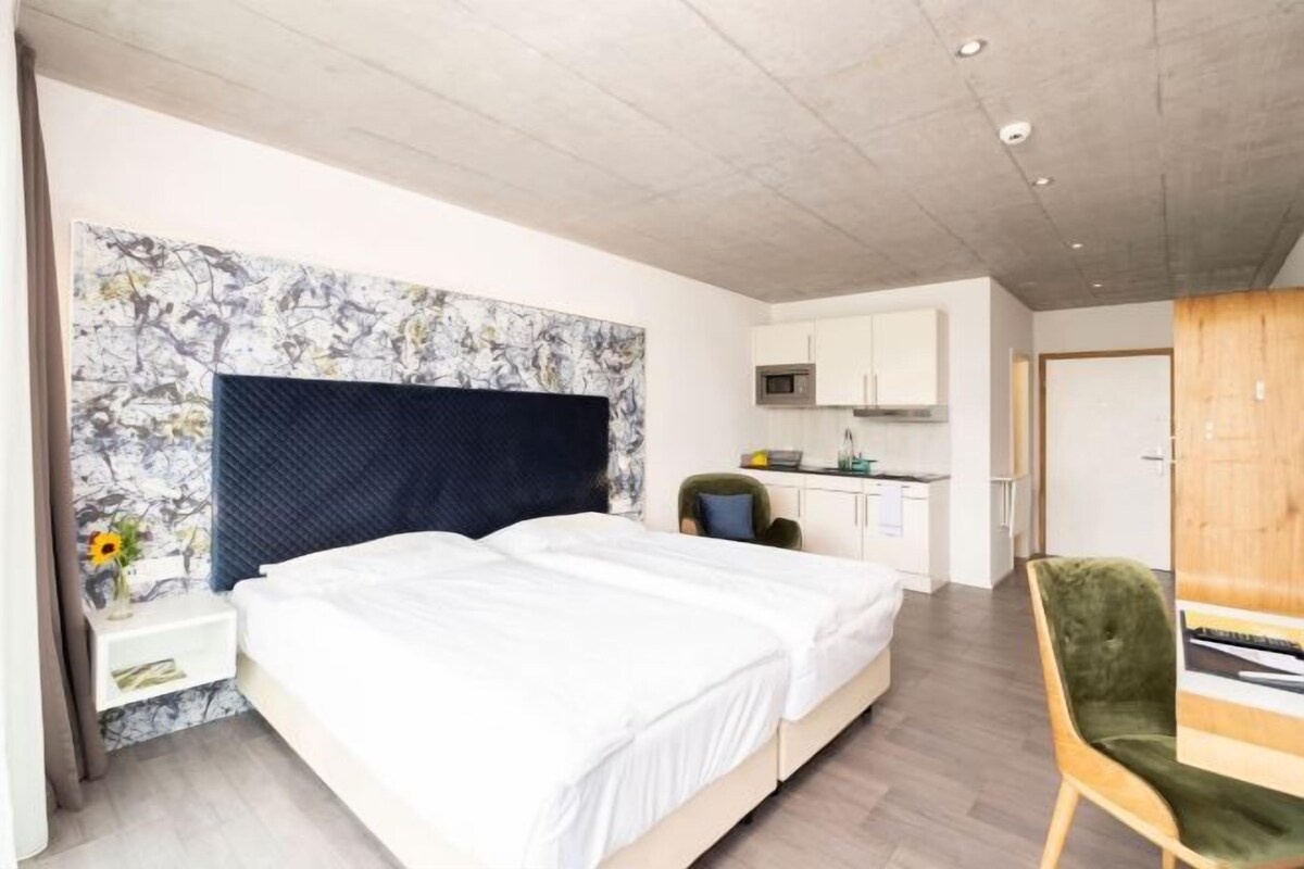 A modern bedroom with a large bed, a desk, and a kitchenette.