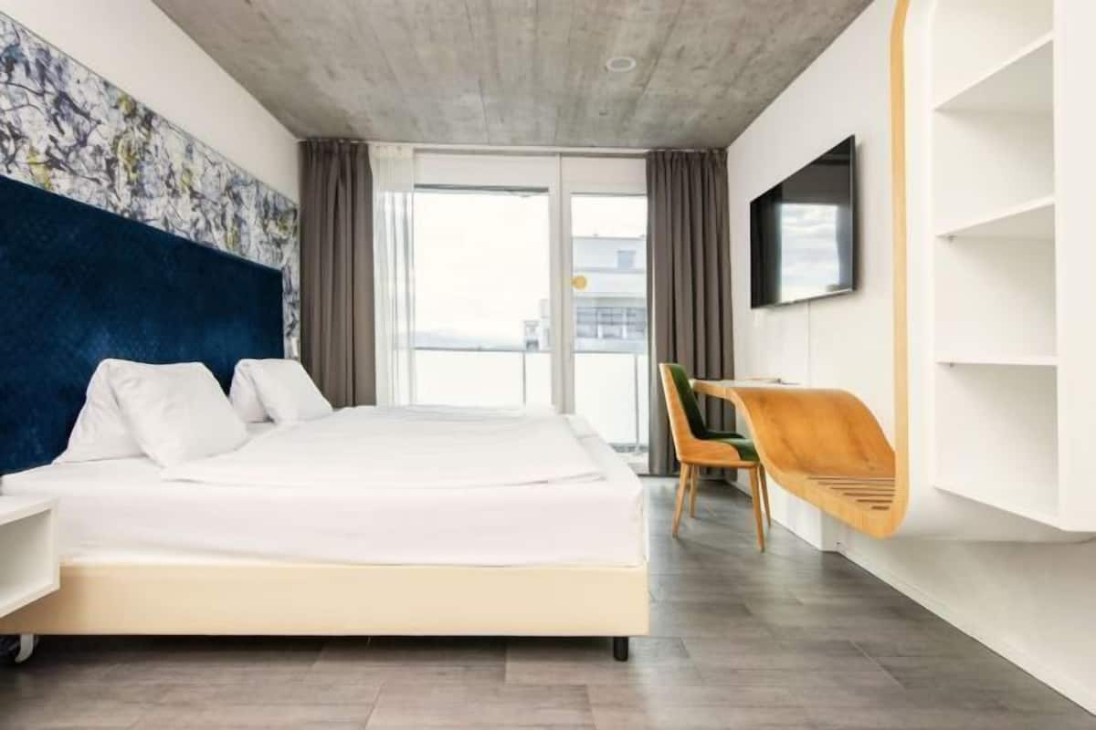 Habitación de hotel moderna con una cama grande, un televisor montado en la pared, una silla de madera y un balcón con vistas.
