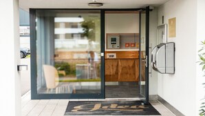 Entrada de hotel con puerta corredera de vidrio, un mostrador de recepción y un pequeño espacio al aire libre visible a través de la puerta.