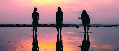 Três silhuetas de pessoas em pé sobre uma superfície refletora, provavelmente uma praia, com um pôr do sol ao fundo.