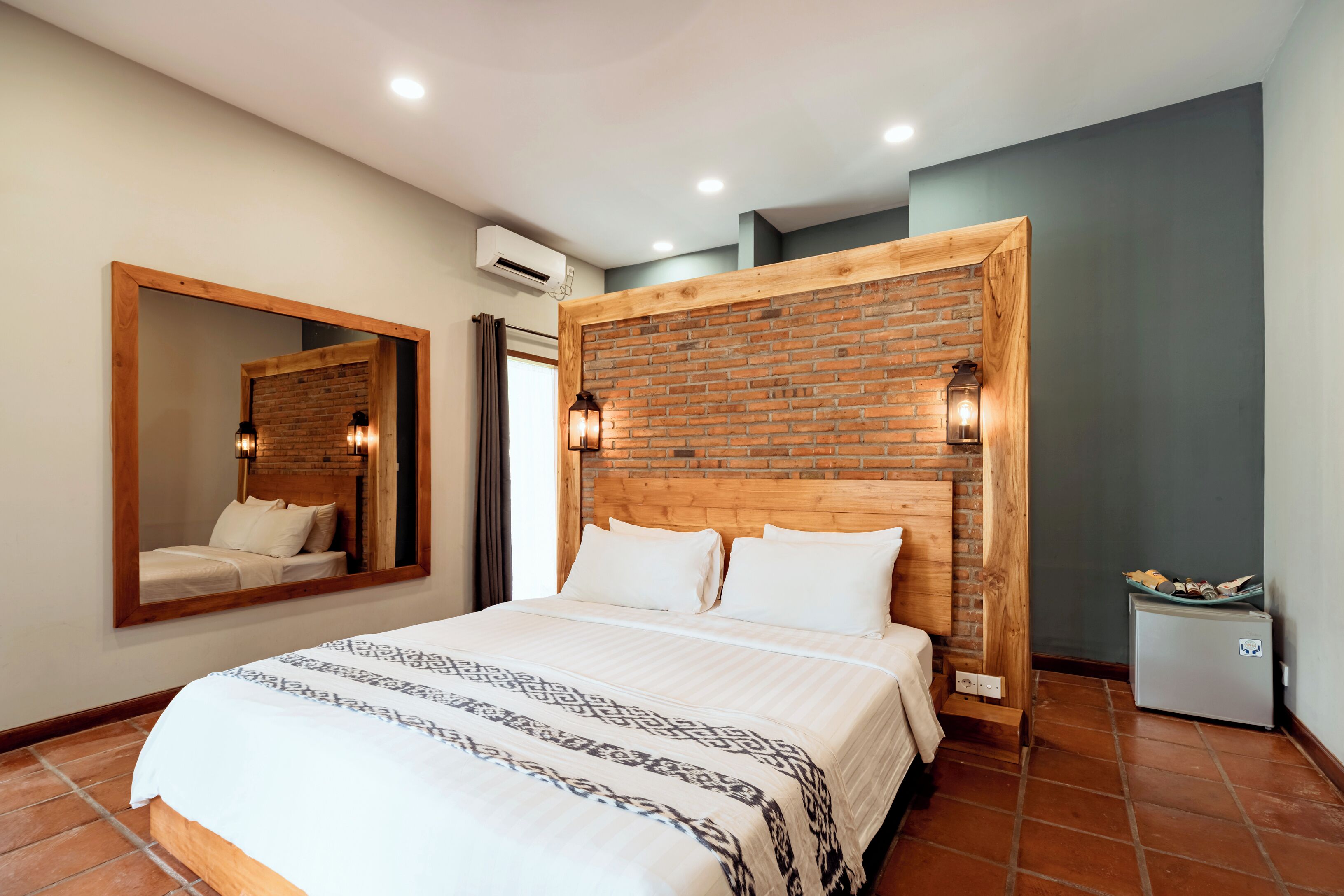 A neatly made bed with a patterned bedspread, a wooden headboard with a brick accent, a small bedside table, and a wall-mounted mirror.