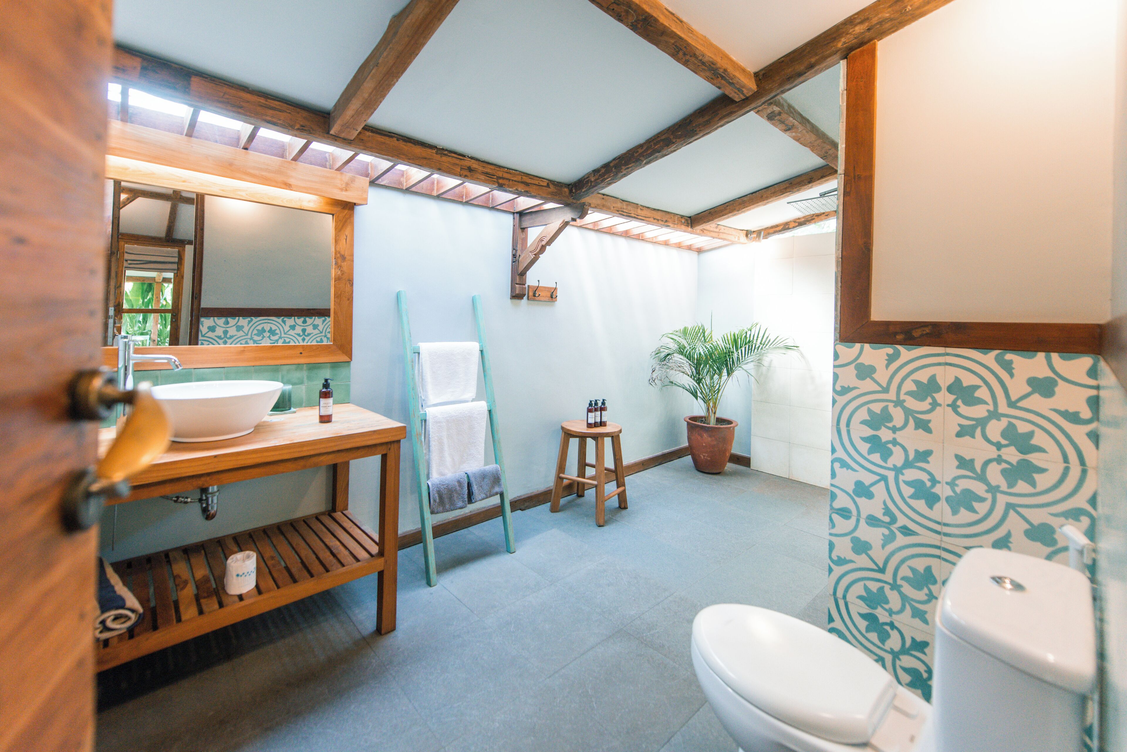 A bathroom with a wooden vanity, a sink, a toilet, a mirror, a towel rack, a small stool, and a potted plant.
