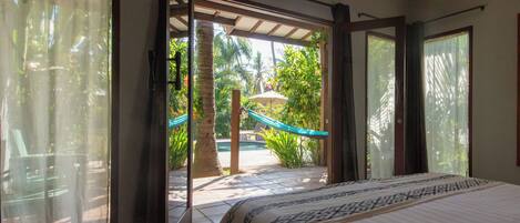 A bedroom with a bed, a view of a pool and palm trees through an open sliding door, and a patio area with a hammock.