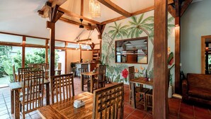 A wooden dining area with tables and chairs, a mural of tropical plants, and a view of greenery outside. - Coconut Garden Resort (Gili Trawangan)
