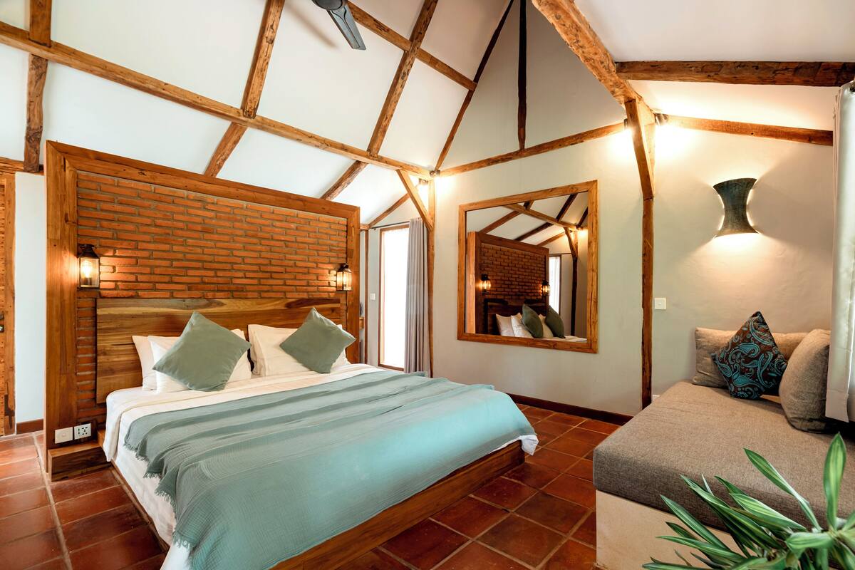 Ein Schlafzimmer mit Holzbalkendecke, einer Ziegelwand als Blickfang, einem Bett mit Kissen, einer Couch mit Polstern und einer Topfpflanze.