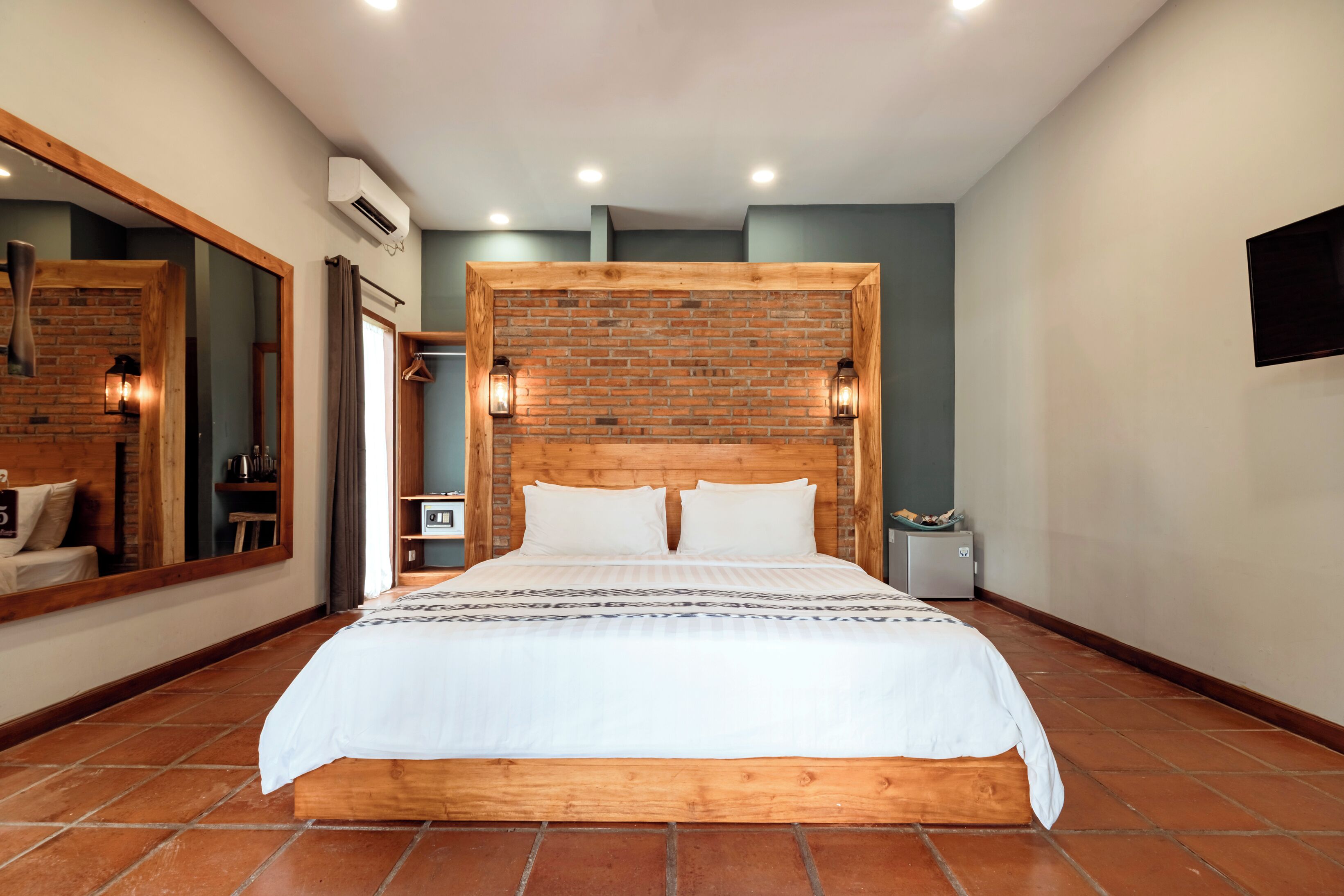 A bedroom with a brick headboard, a large mirror, a bed with white bedding, and a flat-screen TV mounted on the wall.