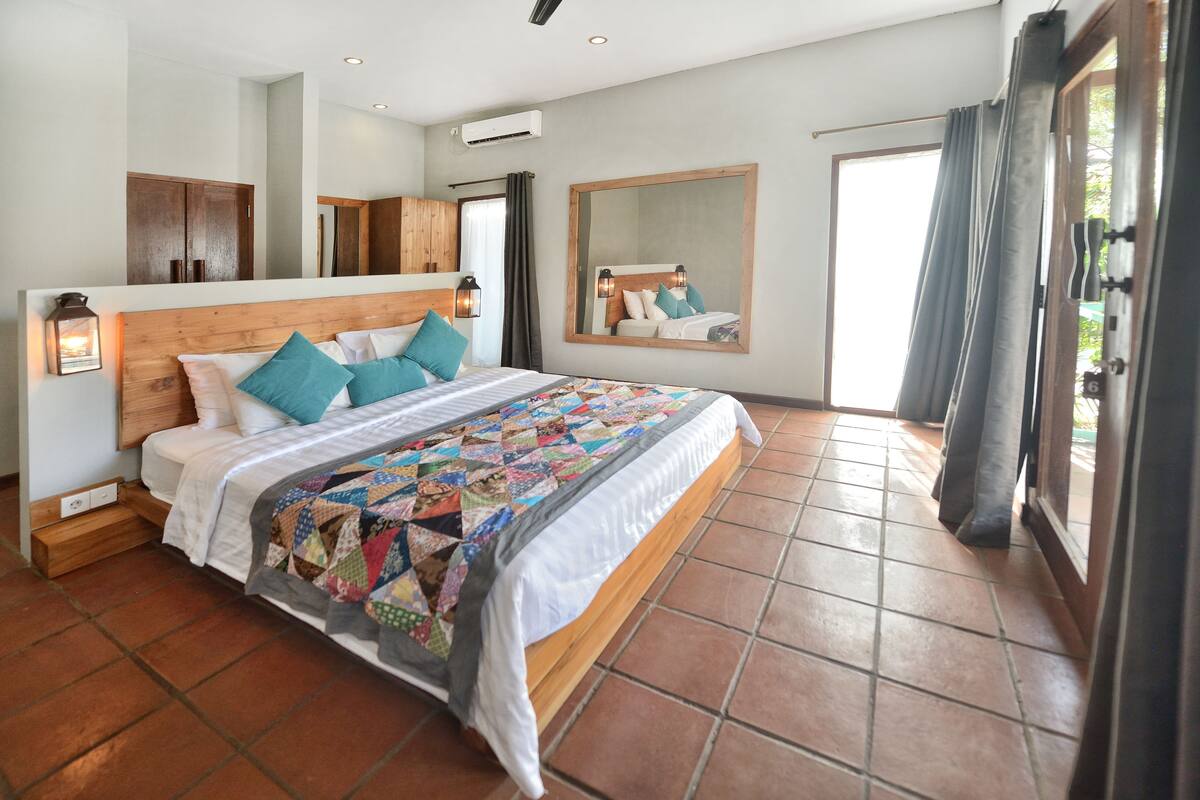 Ein Schlafzimmer mit einem großen Bett, einem Holzkopfteil, einer Steppdecke, einem Spiegel und Blick ins Freie.