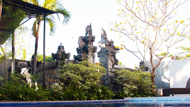 A pool area with stone statues and tropical vegetation. - Putu Guest House 2 (Tibubeneng)