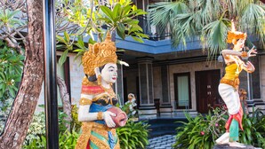A statue of a woman in traditional attire holding a drum, standing on a wooden platform in a courtyard with a building in the background. - Putu Guest House 2 (Tibubeneng)