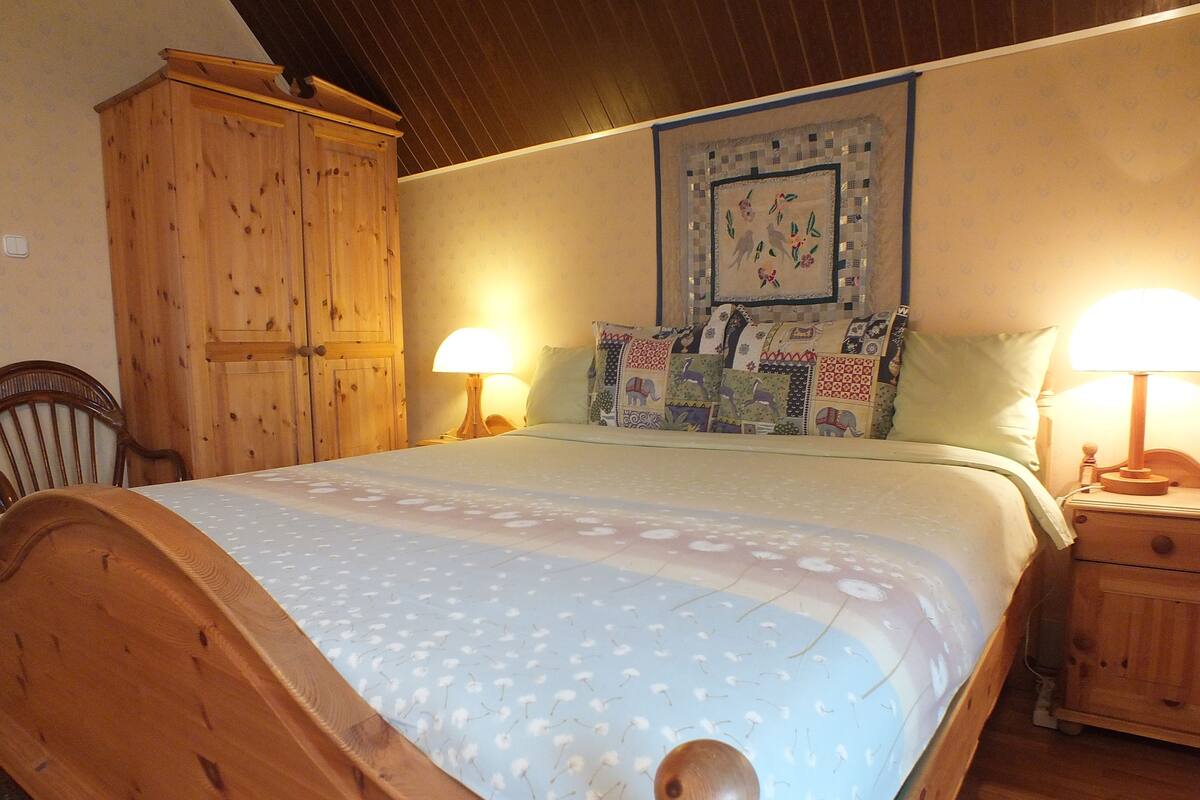 Ein hölzernes Bett mit einem gemusterten Überwurf, zwei Nachttischlampen, ein Holzschrank und ein an der Wand hängendes Bild.