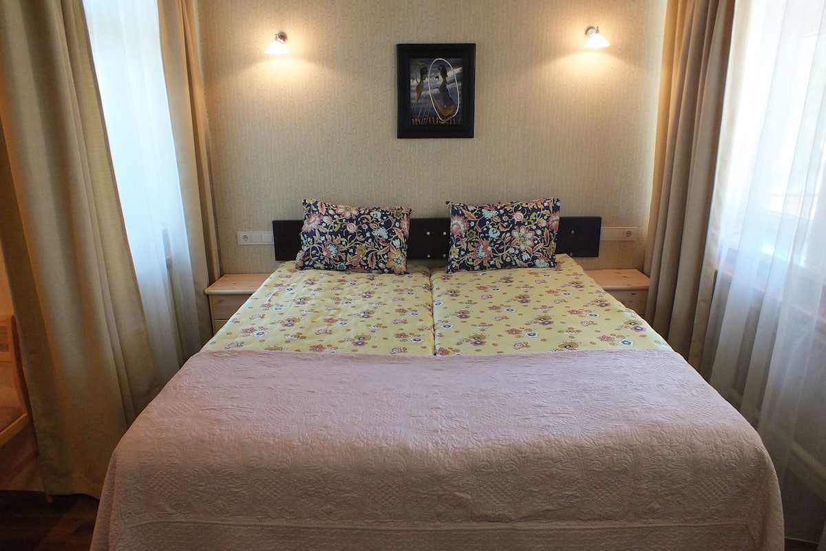 Ein Doppelbett mit Blumenmuster, zwei Nachttische mit Lampen, ein gerahmtes Bild an der Wand und leichte Vorhänge.